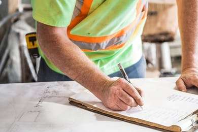 Man in veiligheidskleding schrijft aantekeningen op een notitieboek tijdens een audit, illustratief voor certificeringsprocessen en kwaliteitsmanagement.