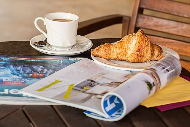 Kopje koffie en croissant op een tafel met een magazine, symboliserend de uitgave en publicatie in de context van kwaliteitsnormen voor de uitgeverssector.