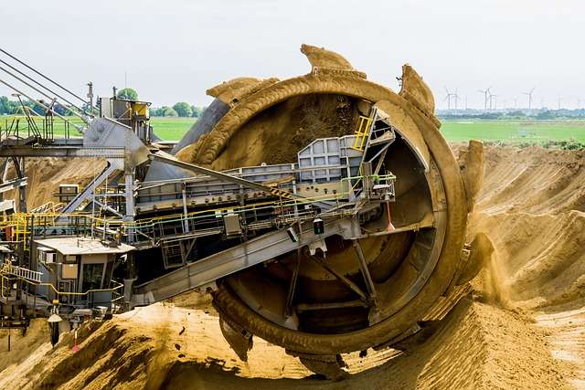 Graafmachine met een grote draaitrommel die zand en grond verplaatst in een mijnbouwomgeving, omringd door groene velden en windturbines op de achtergrond.
