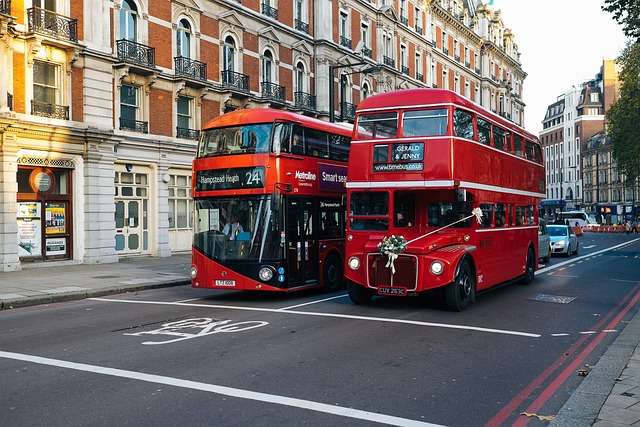 Twee rode bussen in Londen, een moderne dubbeldekker en een klassieke Routemaster, vergezeld door historische gebouwen en een drukke straat.